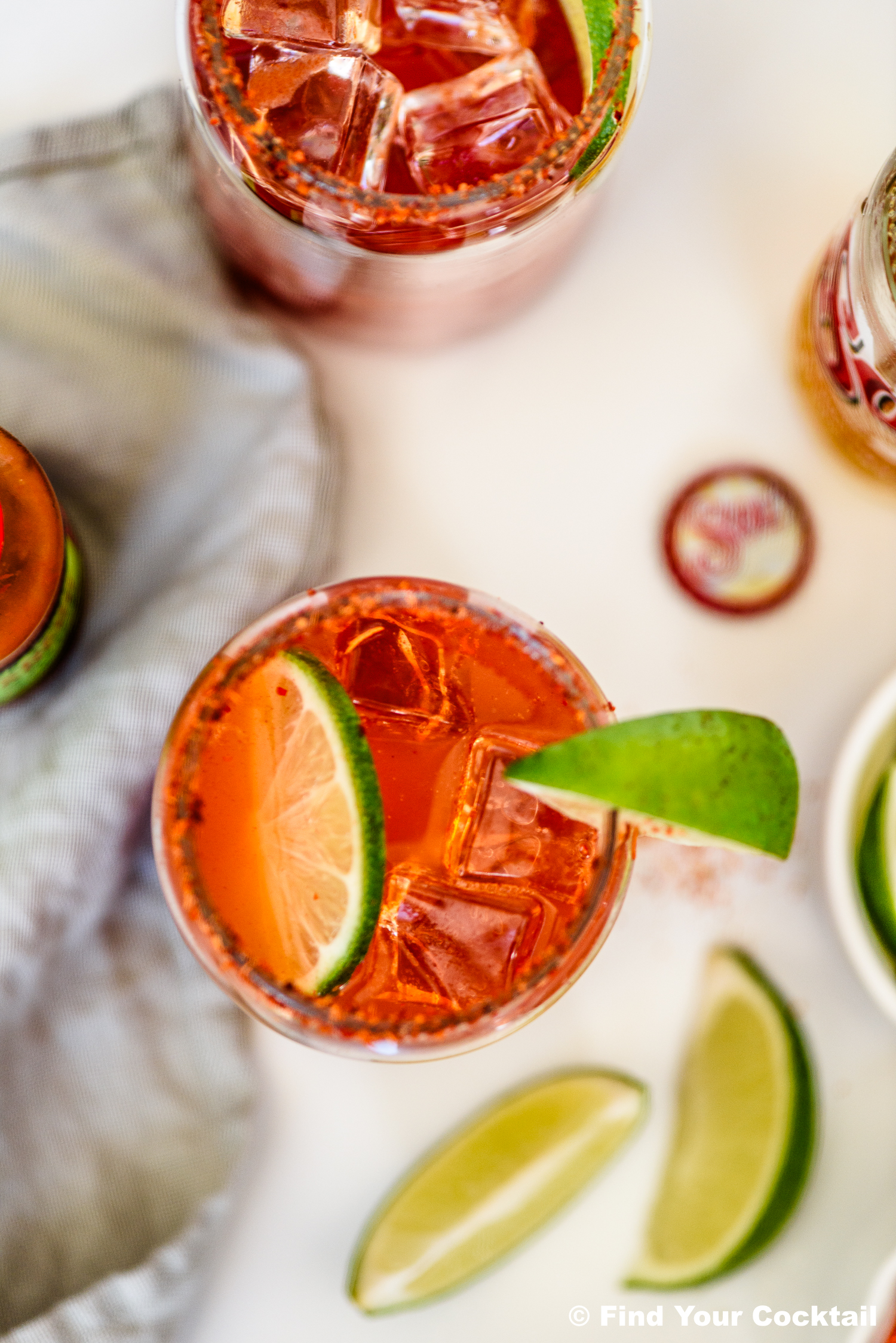 A glass filled with a reddish drink, ice cubes, lime slice, and chili powder rim, next to extra lime wedges and a striped napkin.