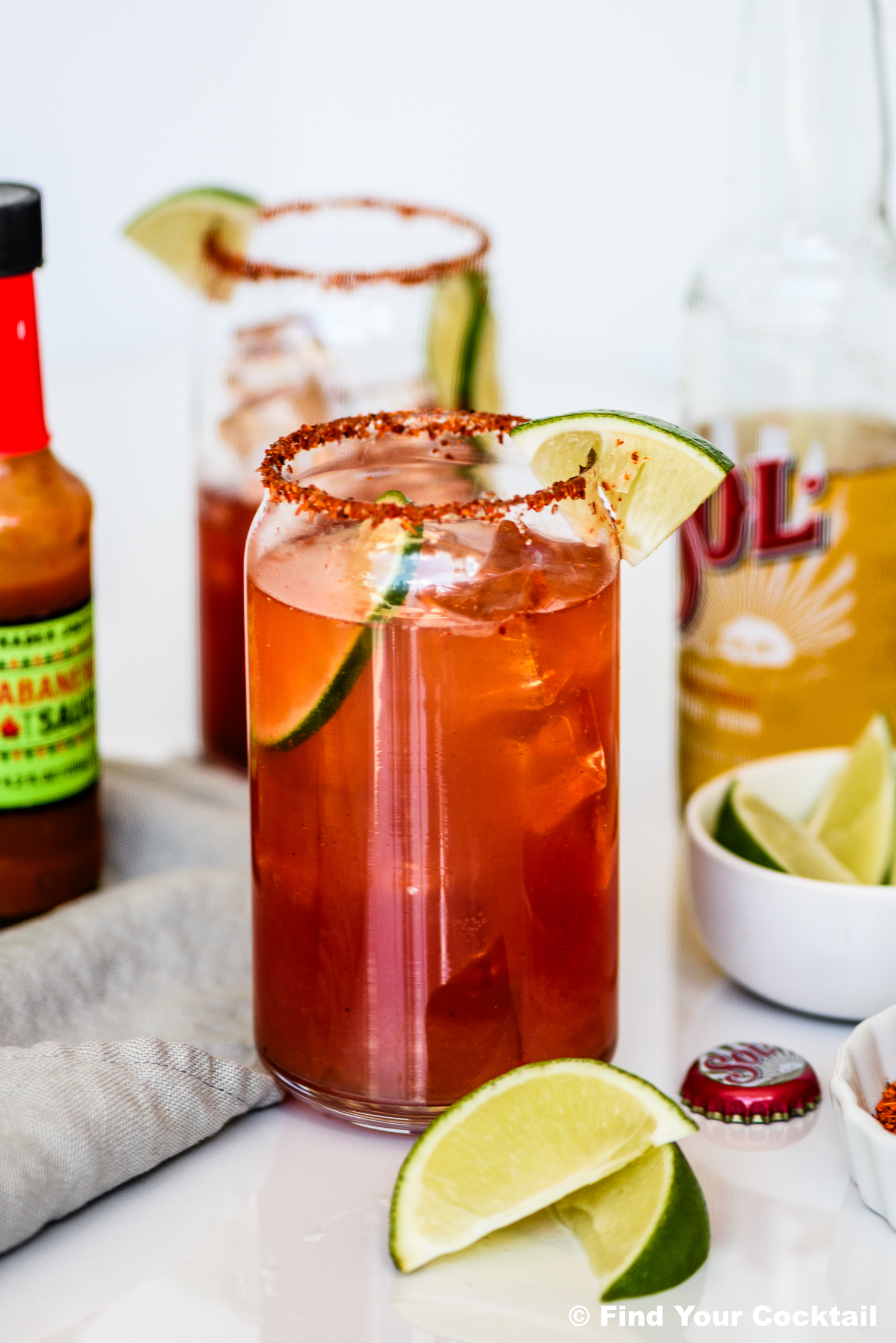 A glass of reddish cocktail with ice, rimmed with chili powder, garnished with lime wedges; hot sauce and a beer bottle are nearby.