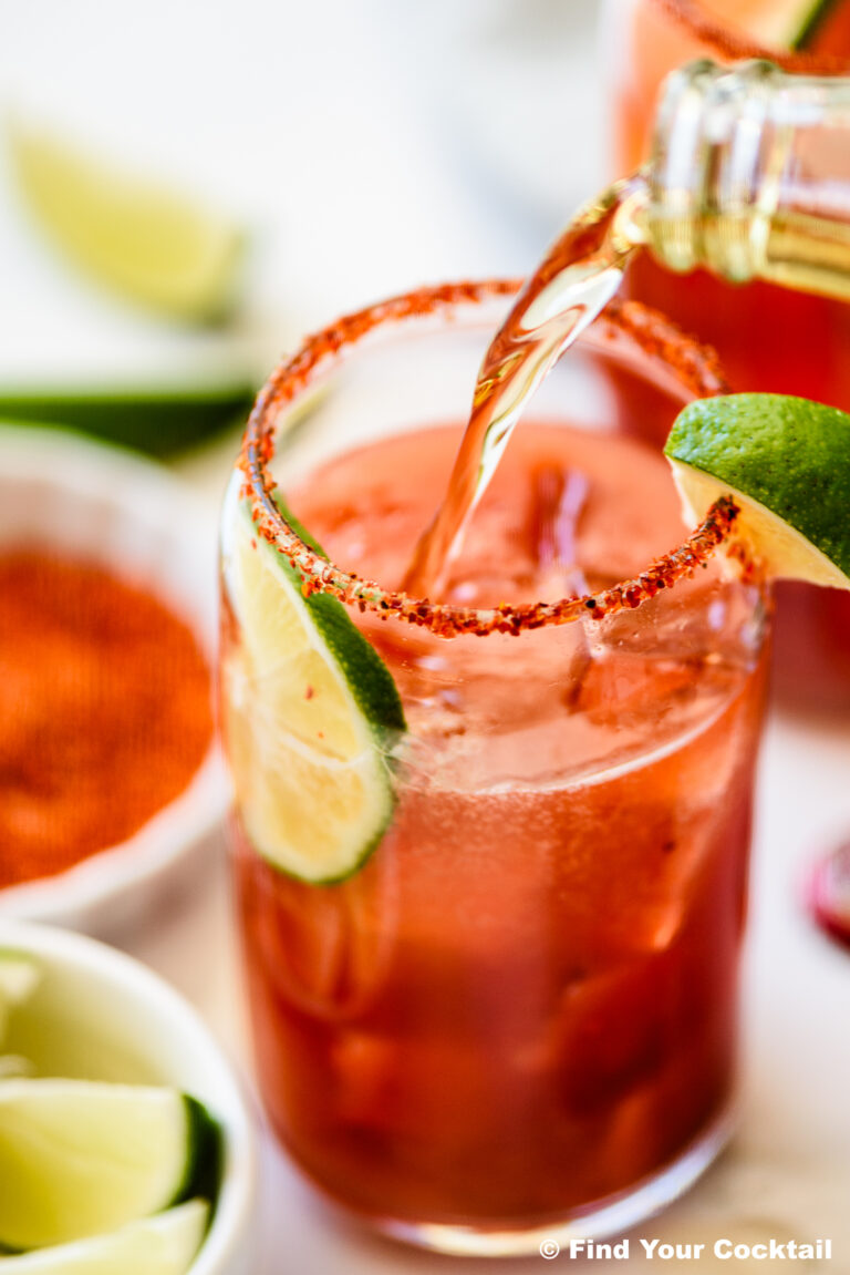 A glass rimmed with chili powder, filled with ice and a reddish drink, is garnished with lime slices while another beverage is being poured in.