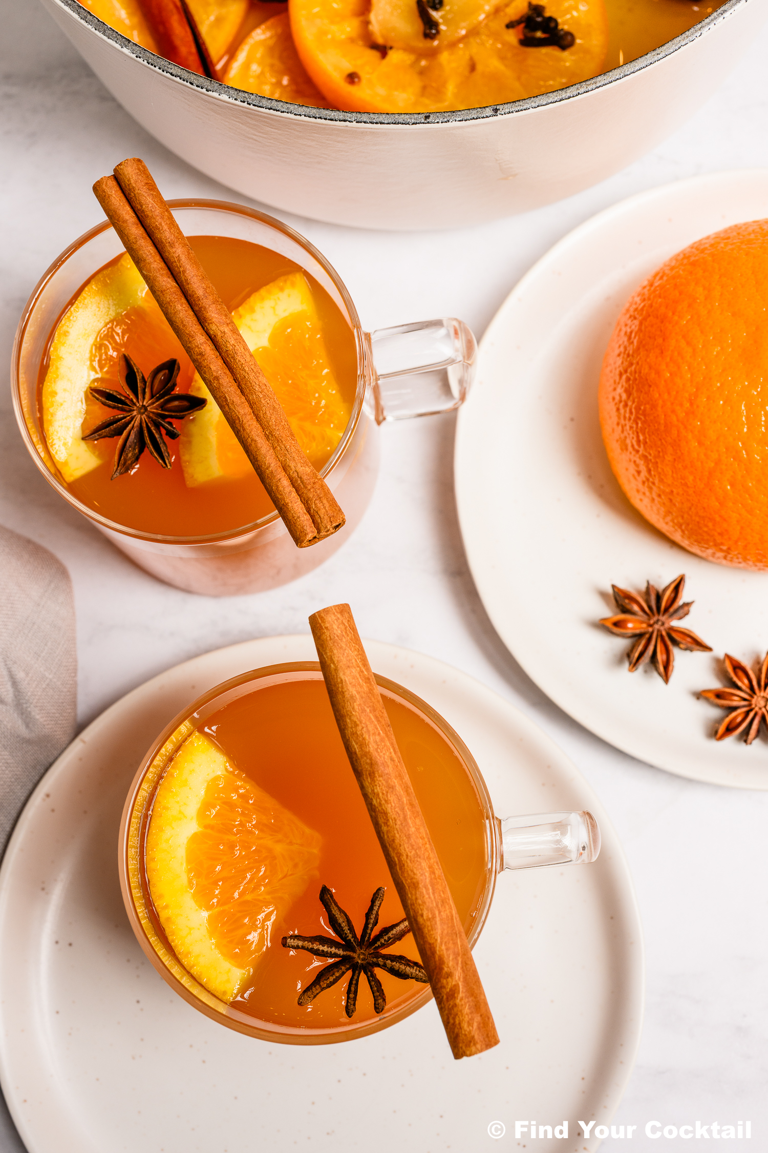 Two glass mugs of orange-hued punch garnished with orange slices, star anise, and cinnamon sticks, next to a whole orange and star anise on a white plate.