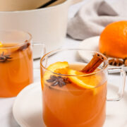 Two clear mugs of orange-hued drink garnished with orange slices, cinnamon sticks, and star anise, with an orange, pot, and ladle in the background.