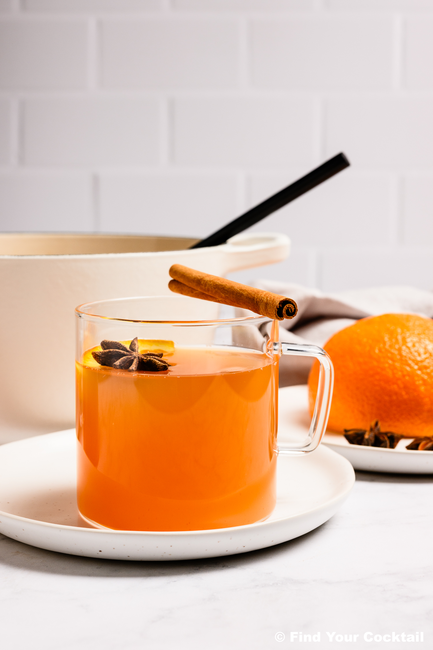 A clear glass mug of orange-colored drink garnished with star anise and a cinnamon stick sits on a white plate next to an orange and a white pot.