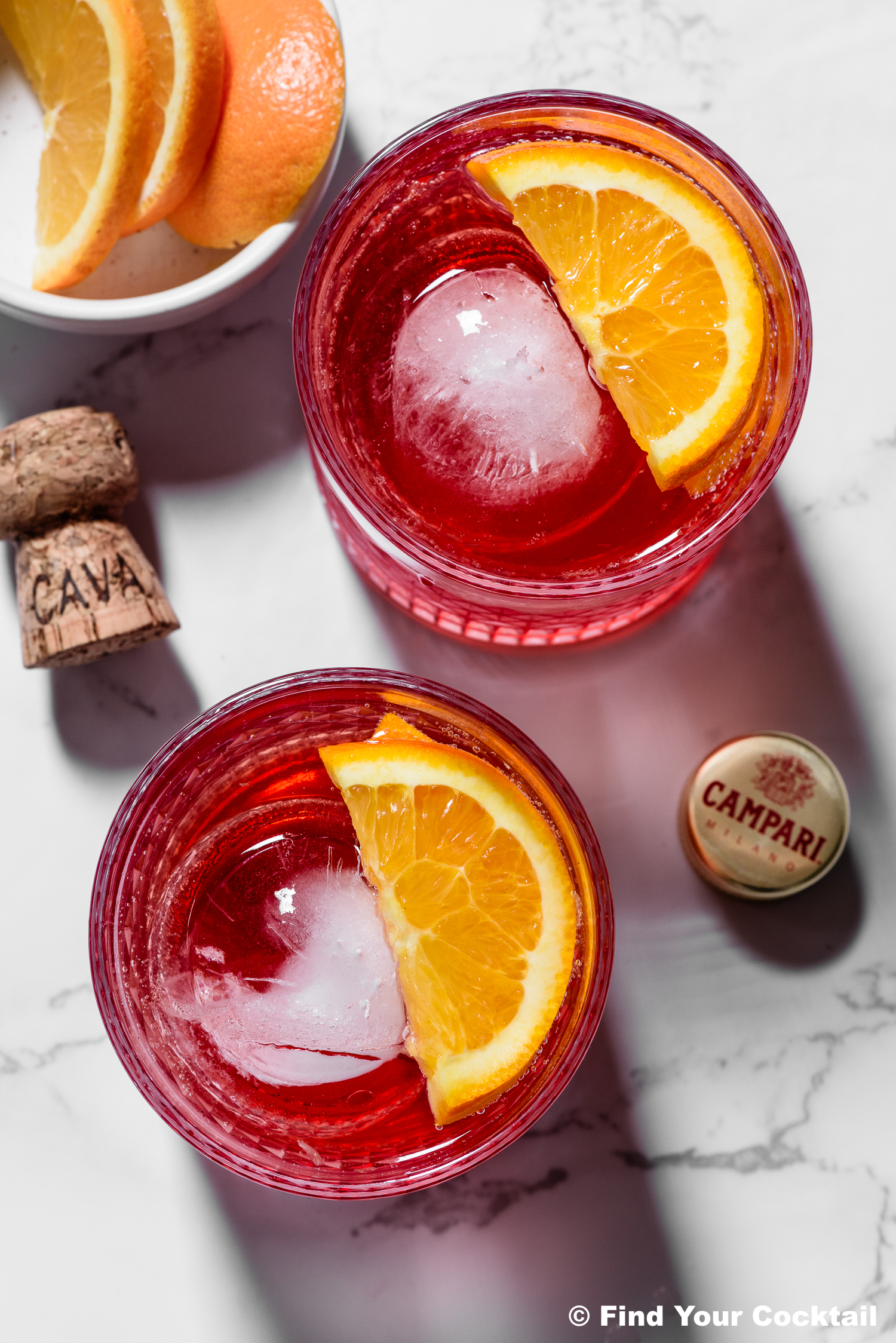 Two glasses of red cocktail with orange wedges and ice spheres on a white surface, next to a bowl of orange slices, a cava cork, and a Campari bottle cap.