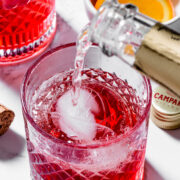 A close-up of a sparkling drink being poured into a glass with ice, next to a bowl of orange slices and a bottle cap on a white surface.