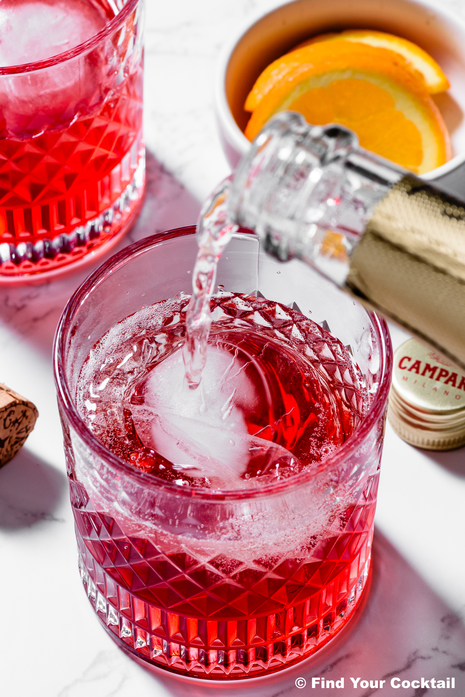 A close-up of a sparkling drink being poured into a glass with ice, next to a bowl of orange slices and a bottle cap on a white surface.