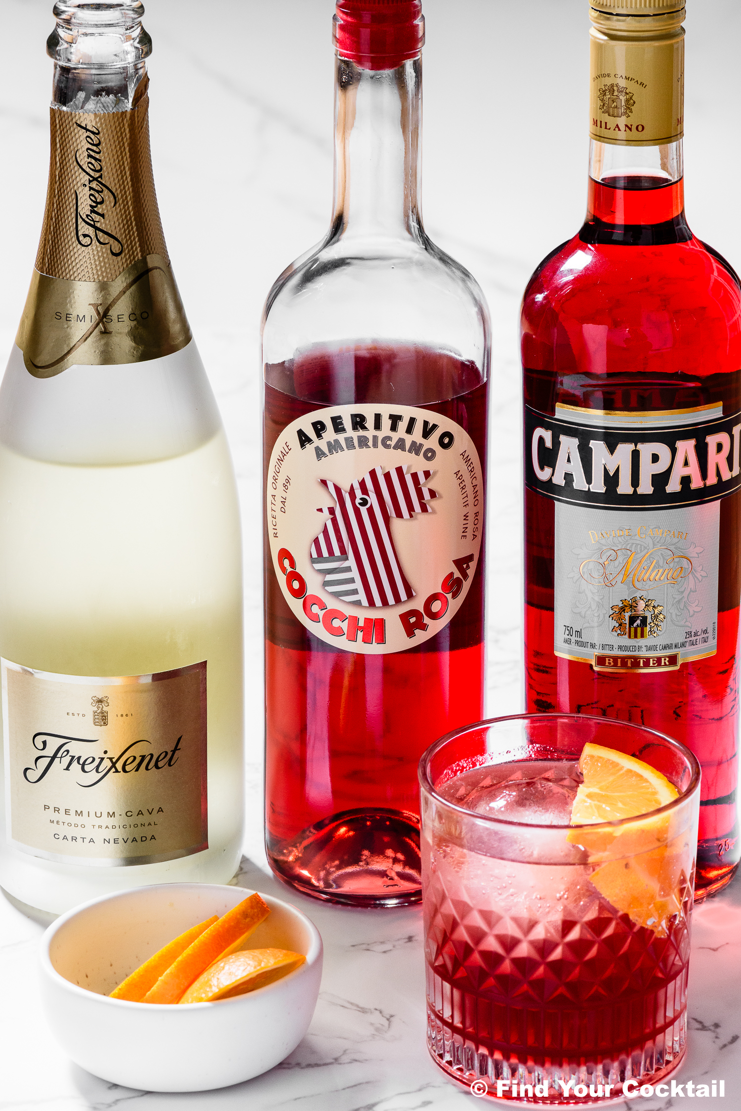 A glass of red cocktail with orange slices is shown beside bottles of Freixenet sparkling wine, Cocchi Rosa aperitif, and Campari, with a small dish of orange wedges.
