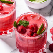 Two glasses of raspberry cocktail with ice, garnished with fresh raspberries, lime wedges, and mint, sit on a marble surface next to a bowl of limes.