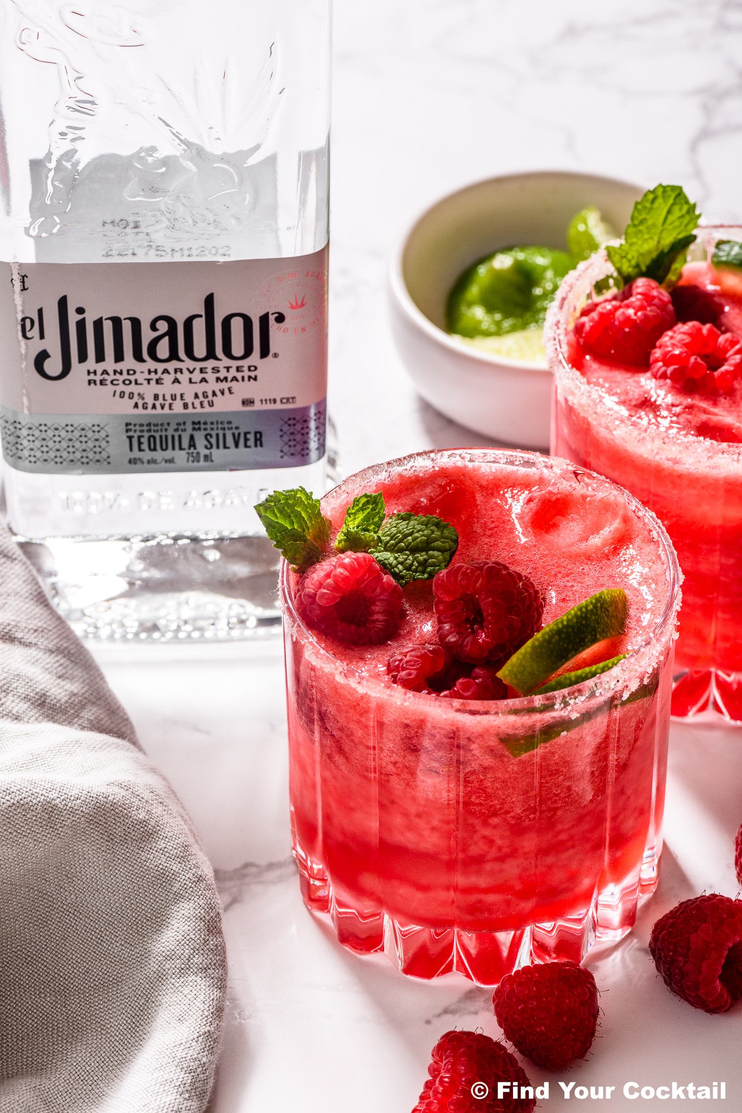 Two glasses of raspberry cocktails garnished with mint and fresh raspberries sit next to a bottle of El Jimador tequila and a bowl of lime wedges on a marble surface.