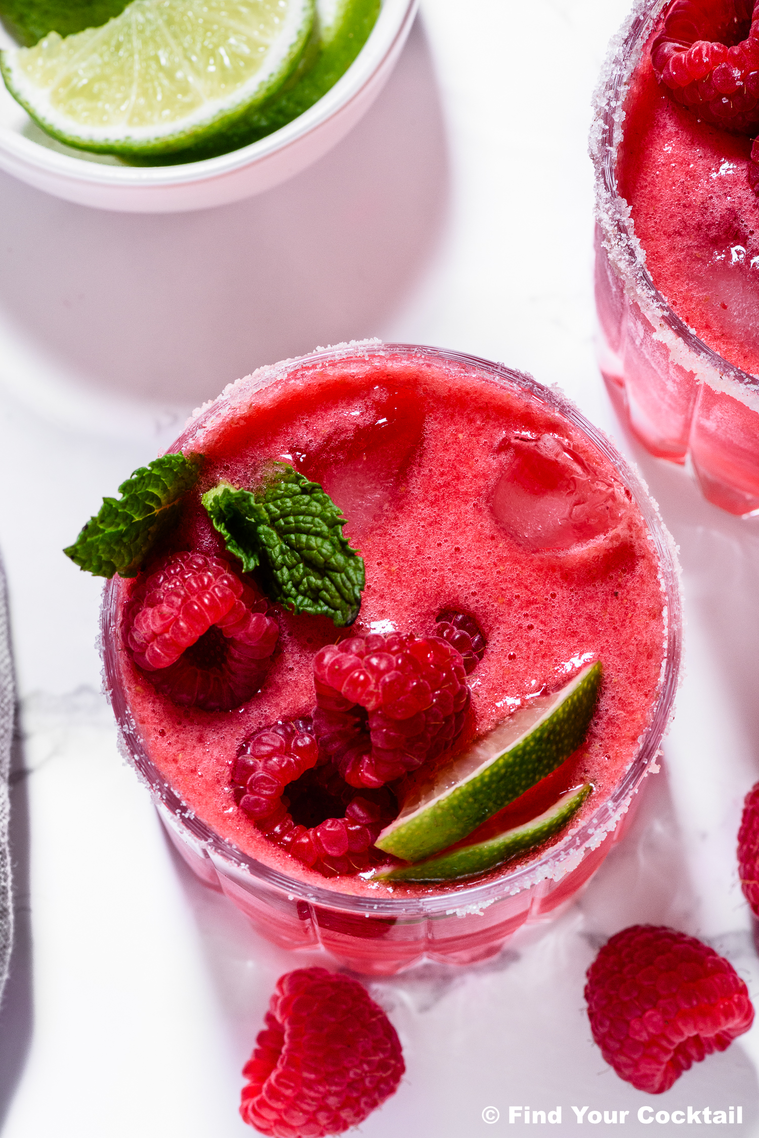 A glass of red raspberry cocktail with ice, garnished with fresh raspberries, mint leaves, and lime wedges. A bowl of lime slices and loose raspberries are nearby.