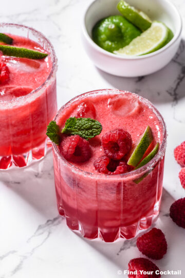 Two glasses of raspberry cocktail with ice, garnished with fresh raspberries, lime wedges, and mint, sit on a marble surface next to a bowl of limes.