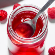 A spoon in a jar of red raspberry syrup, surrounded by fresh raspberries on a white surface.