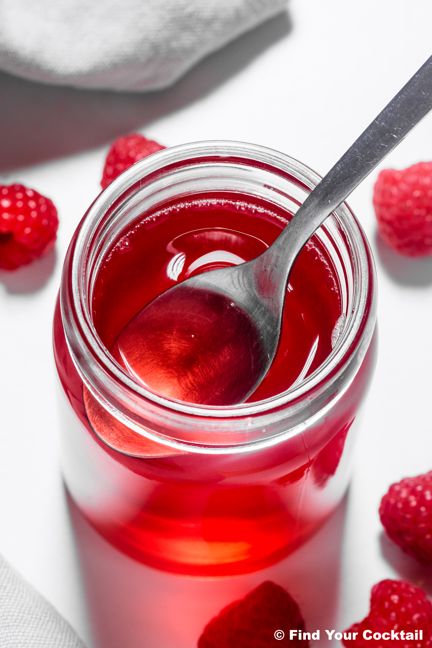 A spoon in a jar of red raspberry syrup, surrounded by fresh raspberries on a white surface.