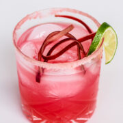 A pink cocktail with ice, garnished with rhubarb strips and a lime wedge, served in a glass with a pink sugar rim.