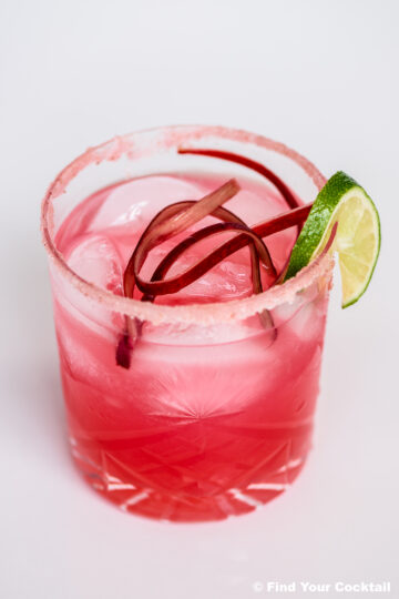 A pink cocktail with ice, garnished with rhubarb strips and a lime wedge, served in a glass with a pink sugar rim.