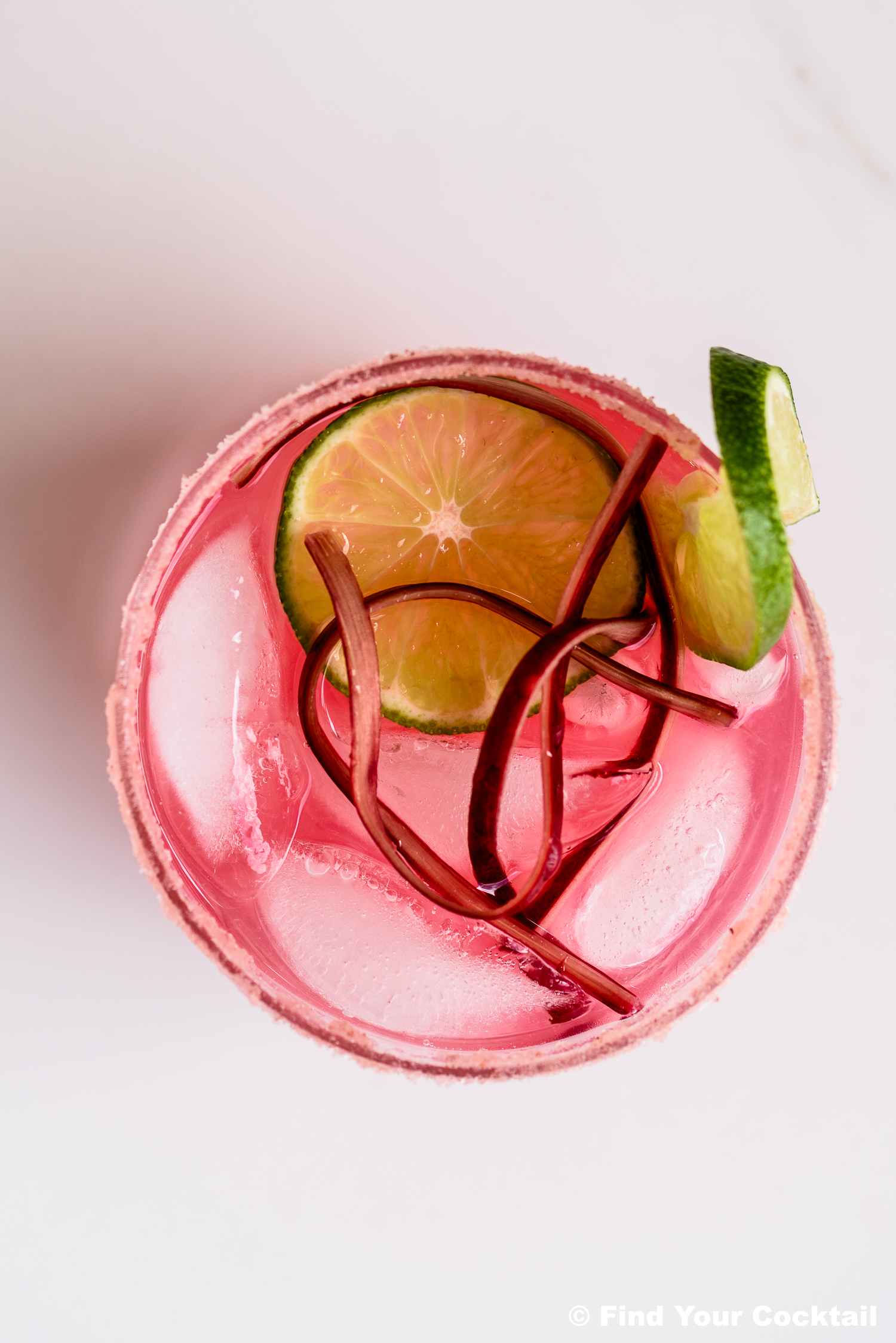A pink cocktail with ice, garnished with two lime slices and thin strips of rhubarb, viewed from above.