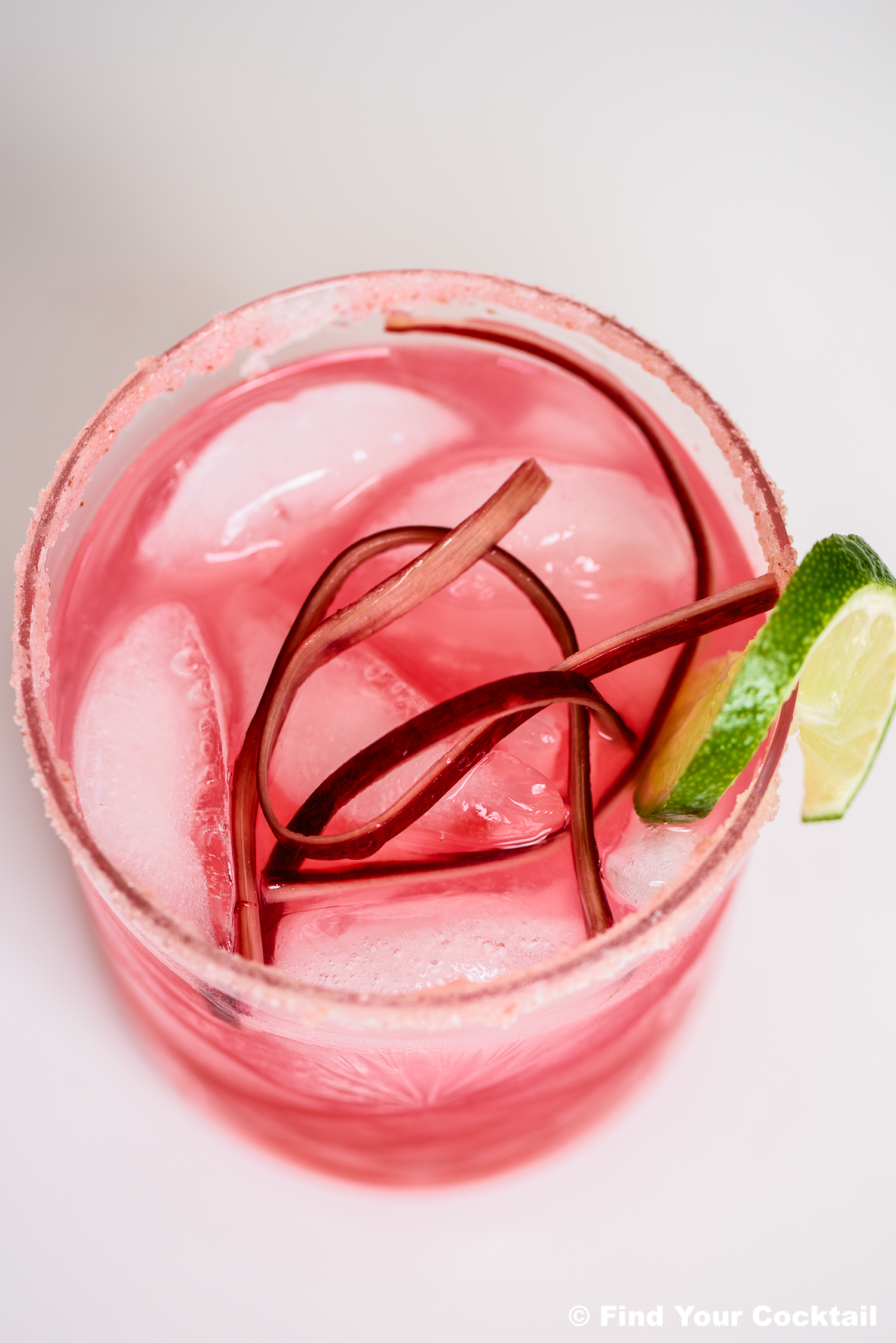 A pink cocktail with ice, garnished with twisted rhubarb strips and a lime wedge, served in a glass with a sugared rim.