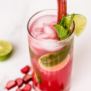 A tall glass of pink cocktail with ice, mint, lime slice, and a rhubarb stalk, garnished with mint and lime; rhubarb pieces and mint leaves are beside the glass.
