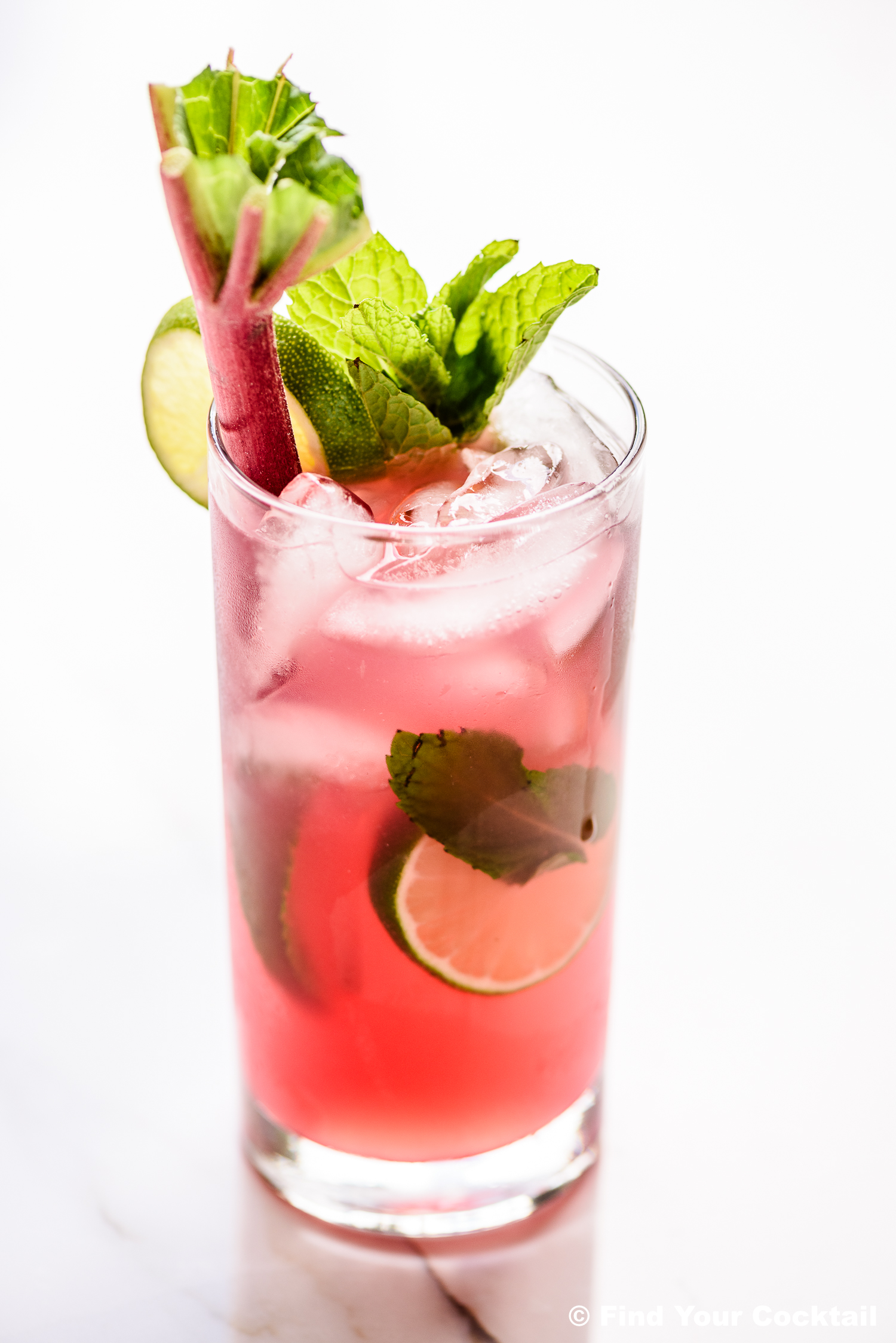 A tall glass of pink cocktail with ice, lime slices, mint leaves, and a rhubarb stalk garnish, on a white surface.