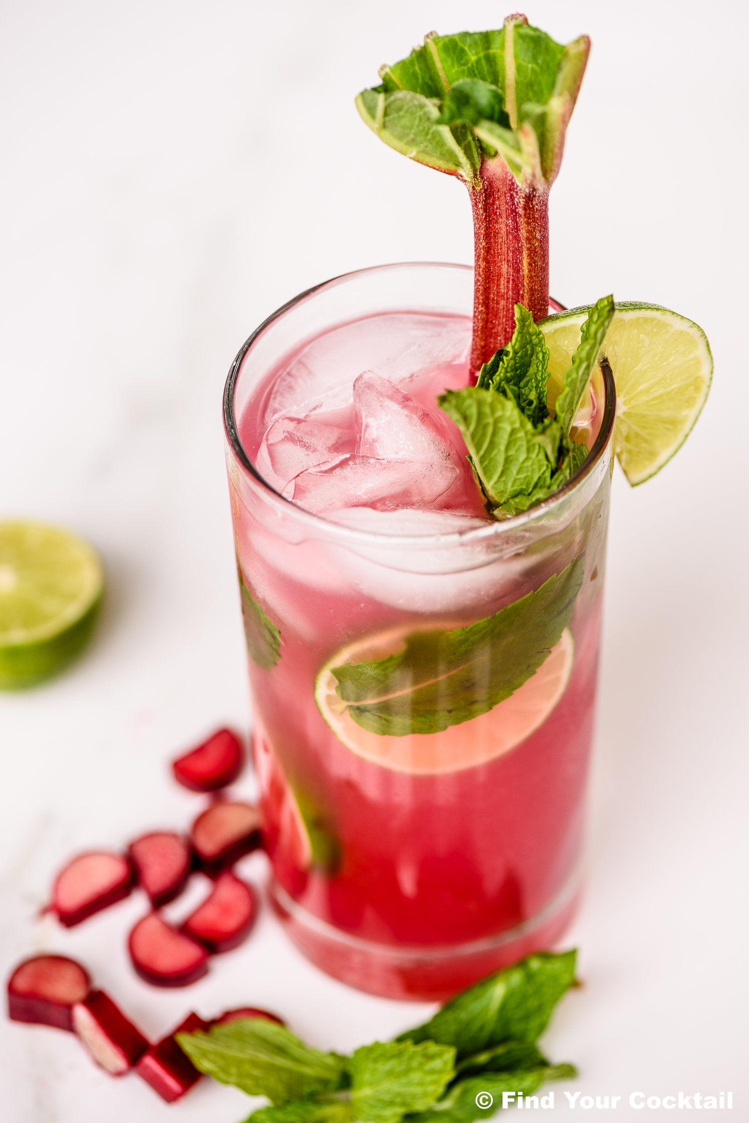 A tall glass of pink cocktail with ice, mint, lime slice, and a rhubarb stalk, garnished with mint and lime; rhubarb pieces and mint leaves are beside the glass.