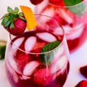 Two glasses of berry cocktail with ice, garnished with fresh strawberries, raspberries, mint leaves, and an orange twist.