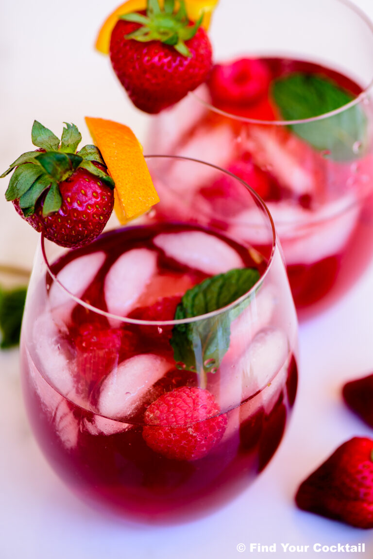 Two glasses of berry cocktail with ice, garnished with fresh strawberries, raspberries, mint leaves, and an orange twist.