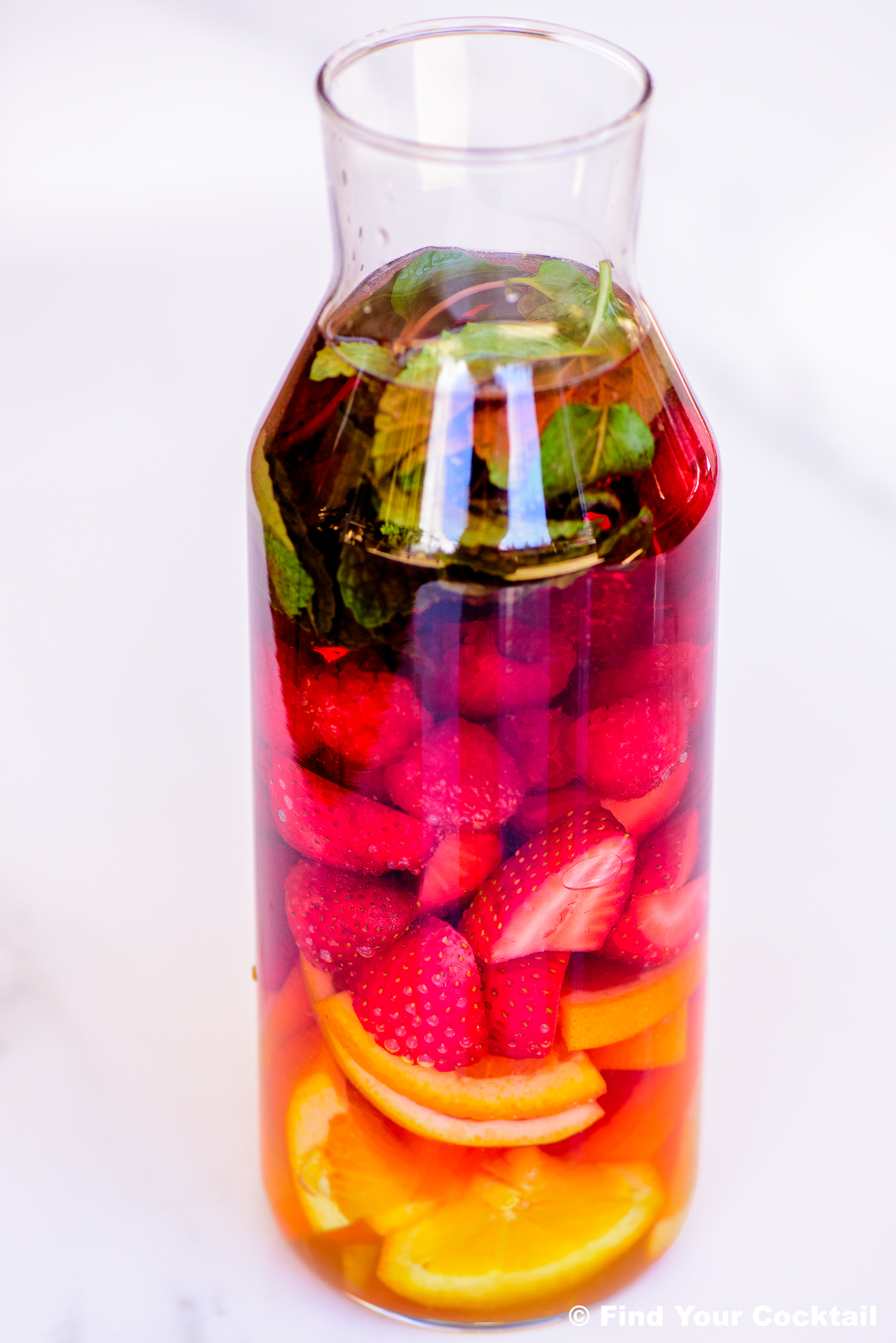 A glass carafe filled with sliced oranges, strawberries, raspberries, mint leaves, and liquid, placed on a white surface.
