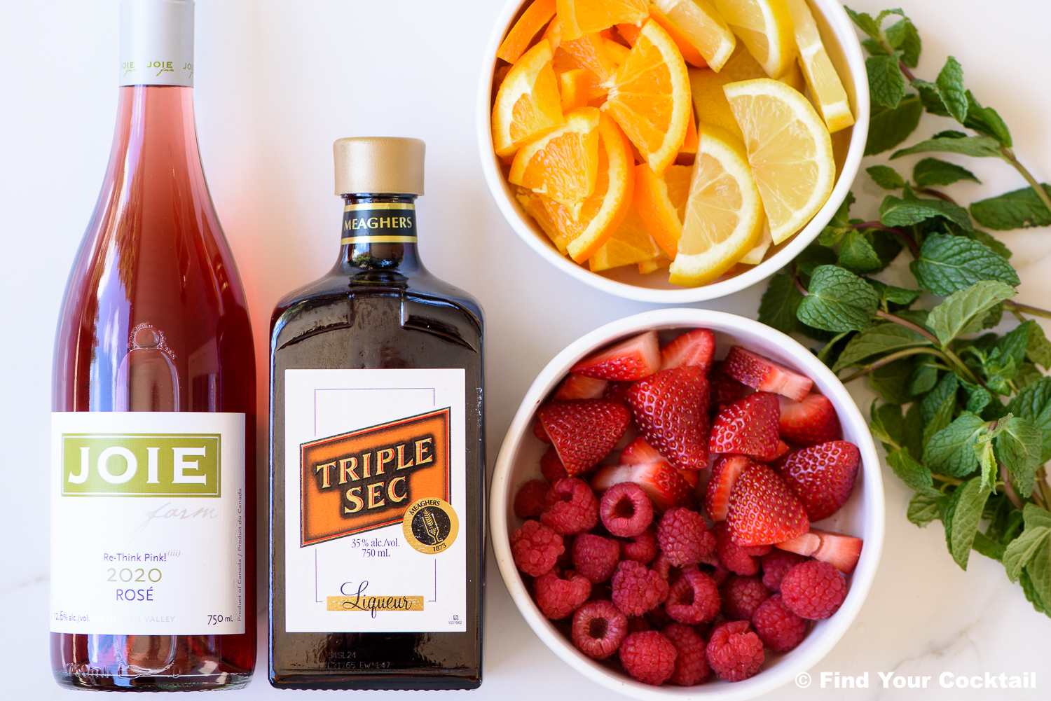 A bottle of rosé wine, a bottle of triple sec, a bowl of sliced citrus, a bowl of mixed berries, and a bunch of fresh mint on a white background.