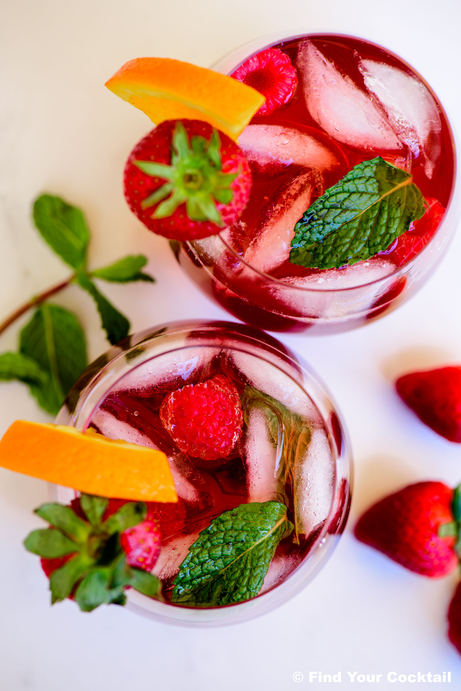 Two glasses of red iced beverage garnished with orange slices, strawberries, raspberries, and fresh mint leaves, placed on a white surface.