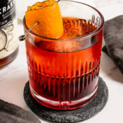 A glass of red cocktail with an orange peel garnish sits on a coaster, next to a bottle of rum, a Campari cap, and a dark cloth napkin.
