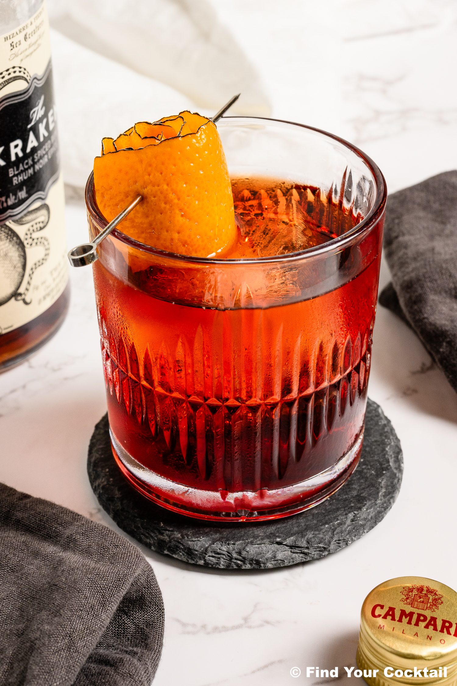 A glass of red cocktail with an orange peel garnish sits on a coaster, next to a bottle of rum, a Campari cap, and a dark cloth napkin.