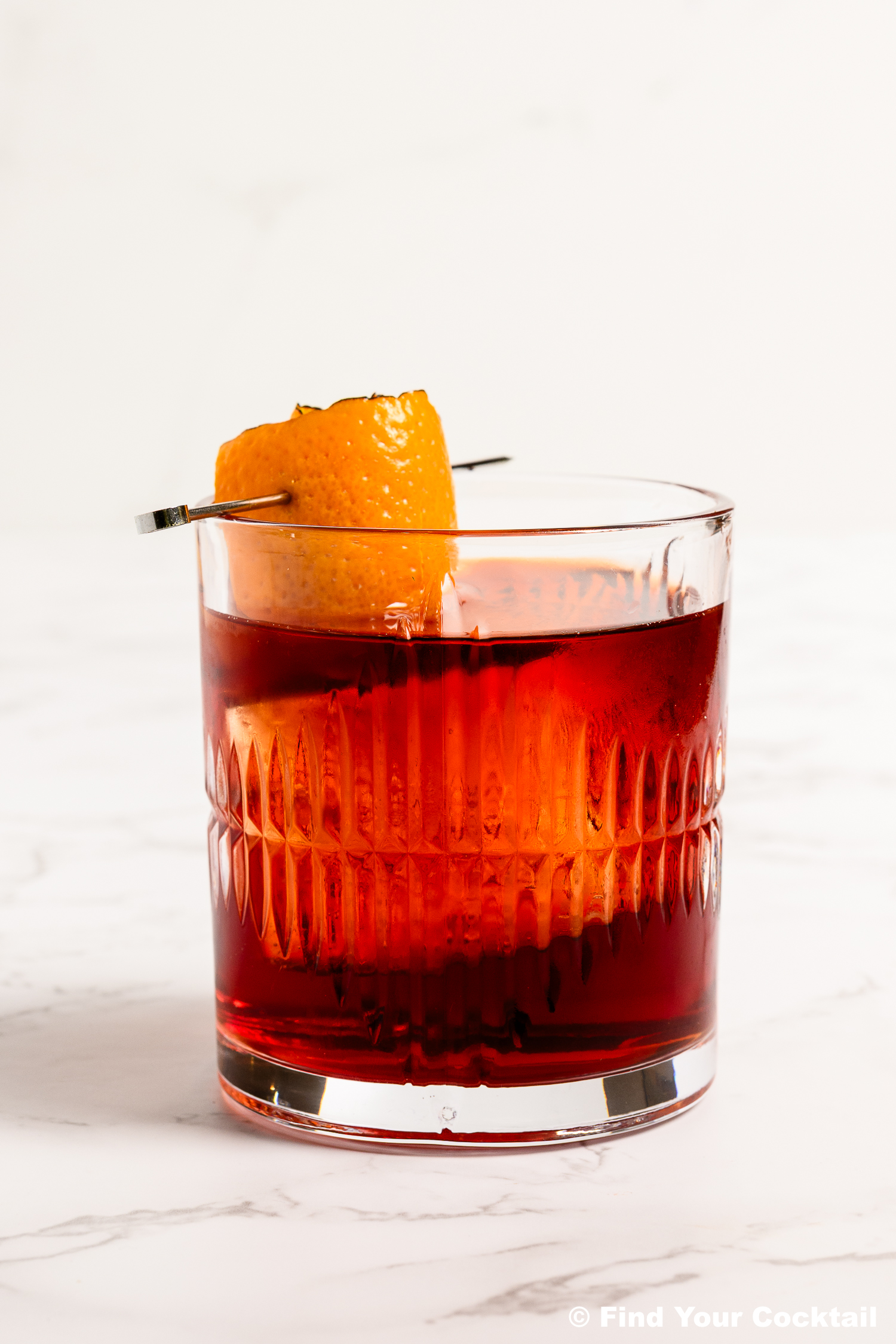 A glass of red cocktail garnished with an orange peel, served on a white marble surface.