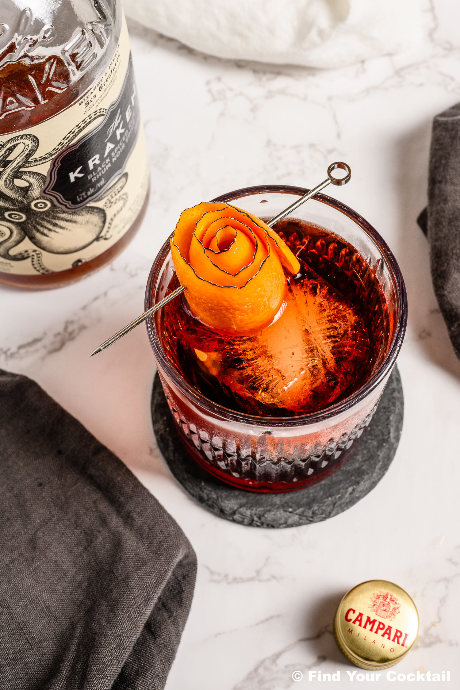 A glass of cocktail with a large ice cube, garnished with an orange peel rose on a metal pick. Kraken rum bottle, Campari cap, and dark napkins are nearby on a marble surface.