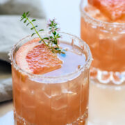 A glass of pink cocktail with ice, garnished with a grapefruit slice, a sprig of thyme, and a salted rim. Another similar drink is in the background.