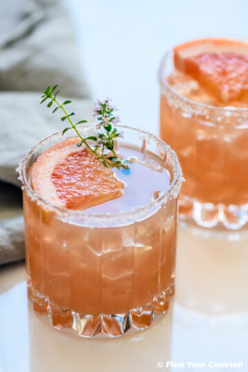 A glass of pink cocktail with ice, garnished with a grapefruit slice, a sprig of thyme, and a salted rim. Another similar drink is in the background.