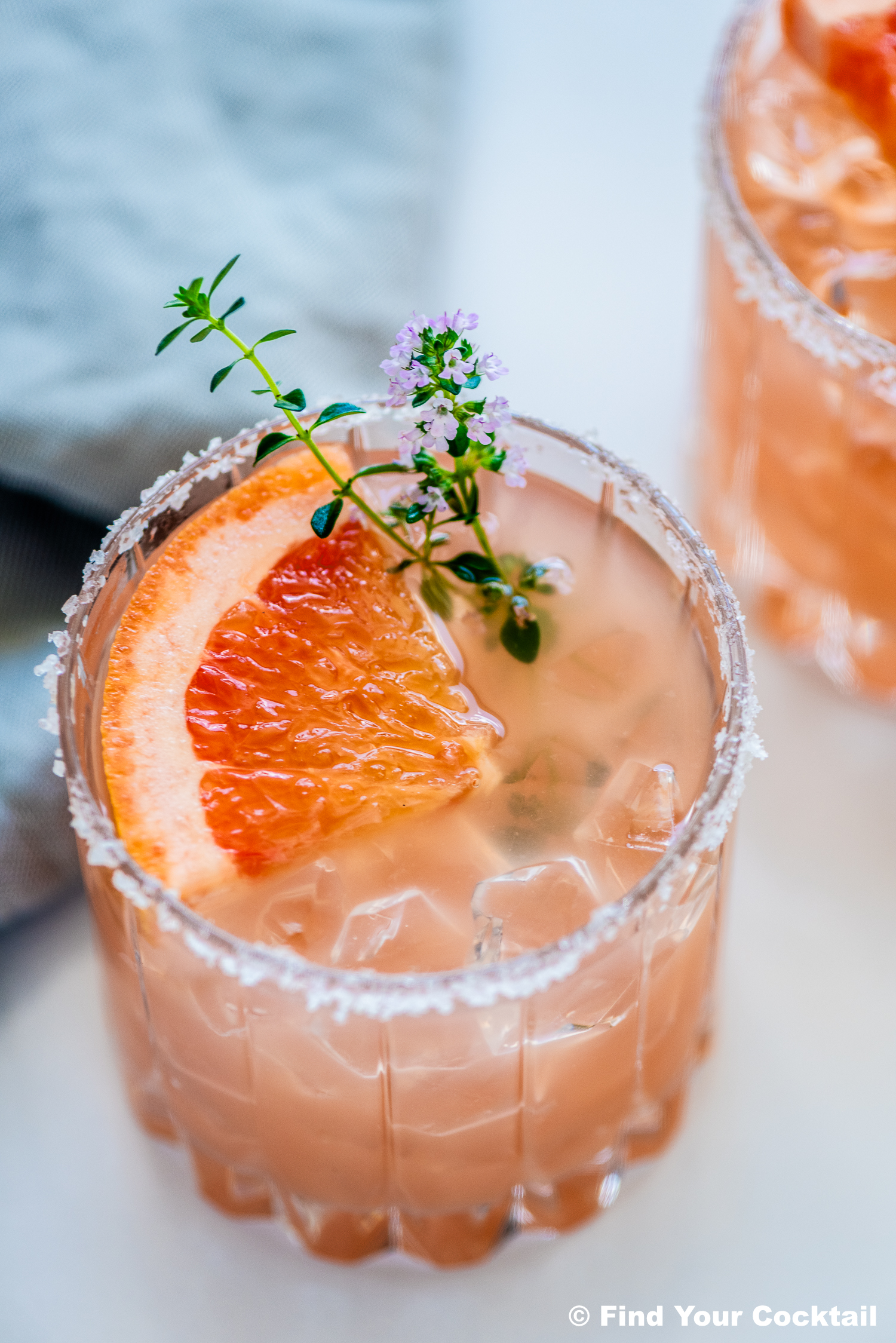A glass of pink cocktail with ice, a salted rim, garnished with a grapefruit slice and a sprig of herbs and flowers.