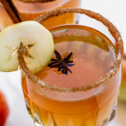 A cocktail in a glass with a brown sugar rim, garnished with a green apple slice, star anise, and a cinnamon stick.