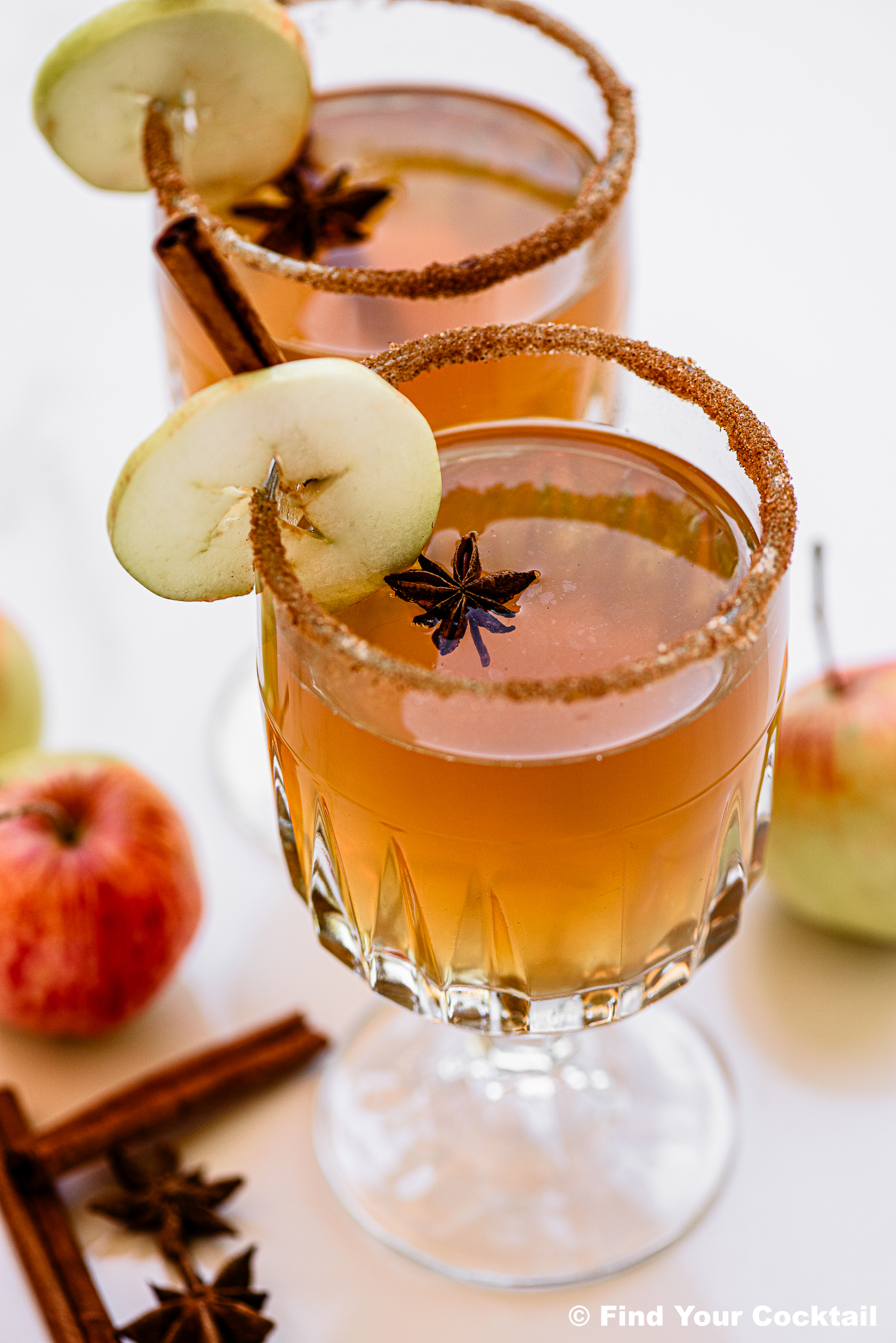 Two glasses of apple cider cocktail garnished with apple slices, star anise, and cinnamon sticks, featuring a brown sugar rim. Apples and spices are placed nearby.