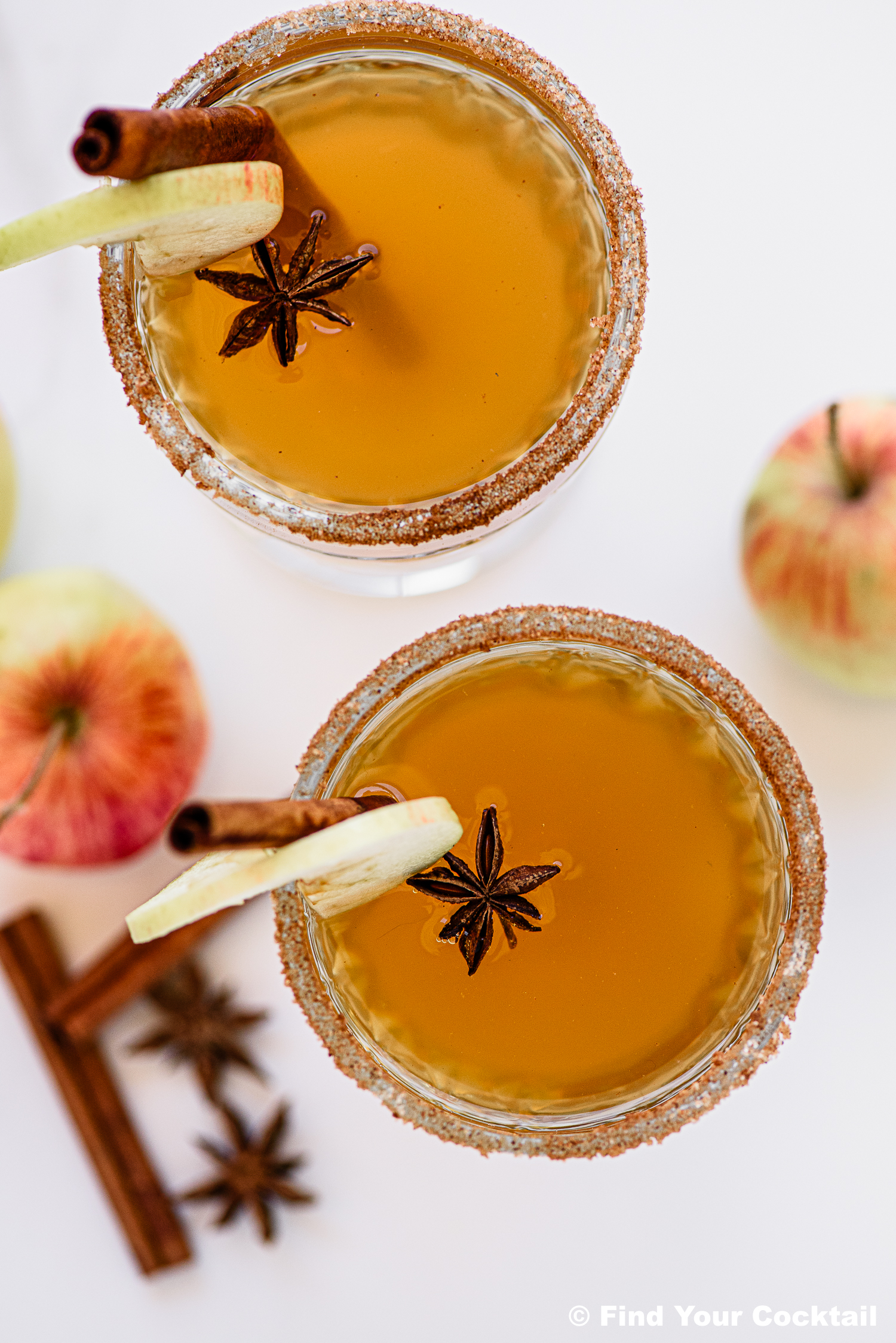 Two apple cider cocktails garnished with apple slices, star anise, and cinnamon sticks, with sugared rims. Apples and spices are on the white surface beside the drinks.