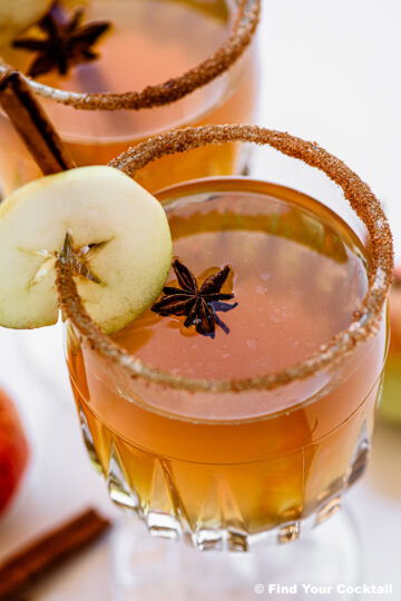 A cocktail in a glass with a brown sugar rim, garnished with a green apple slice, star anise, and a cinnamon stick.