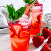 Two glasses of strawberry-infused iced drink with metal straws, garnished with fresh mint and basil, sit on a marble surface next to whole strawberries.