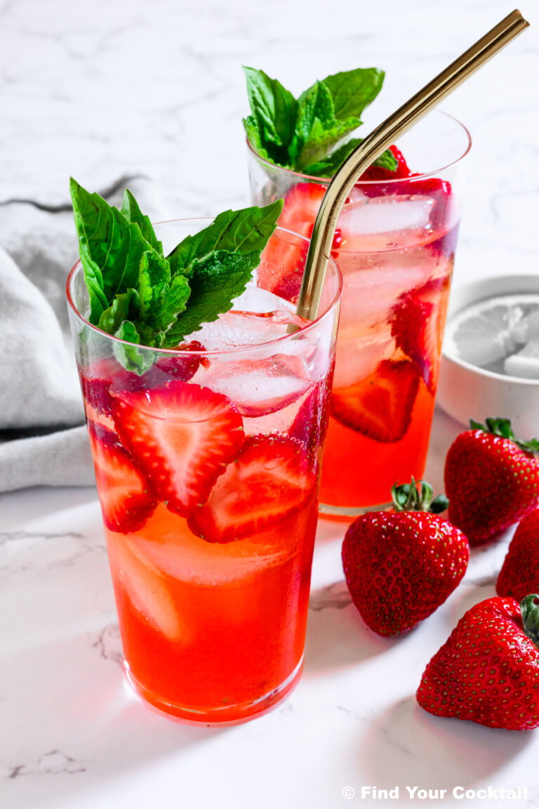 Two glasses of strawberry-infused iced drink with metal straws, garnished with fresh mint and basil, sit on a marble surface next to whole strawberries.
