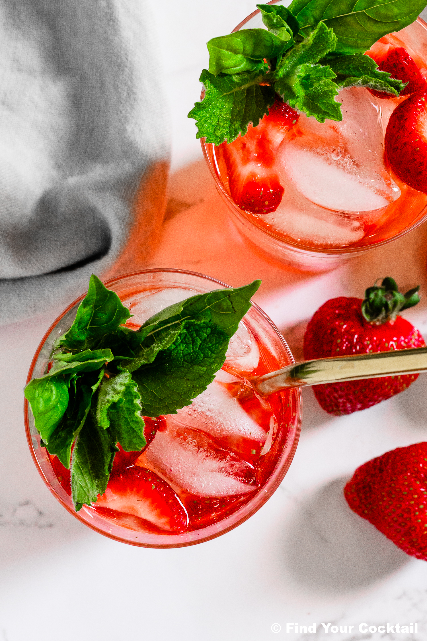 Two glasses of strawberry cocktail with ice, fresh mint garnish, and sliced strawberries on a white surface, next to whole strawberries and a gray cloth.