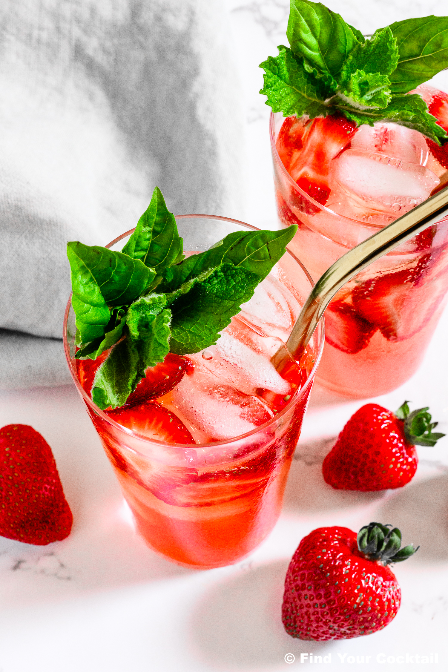 Two glasses of strawberry-infused iced drink with gold straws, garnished with fresh basil leaves, surrounded by whole strawberries on a white surface.