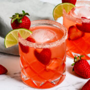 Two glasses of pink cocktail with ice, garnished with strawberries and lime slices, sit on a marble surface with a gray napkin and loose strawberries nearby.