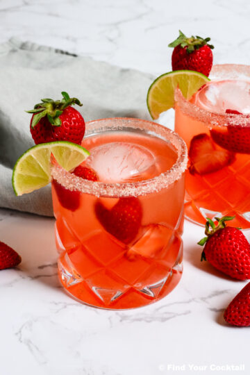 Two glasses of pink cocktail with ice, garnished with strawberries and lime slices, sit on a marble surface with a gray napkin and loose strawberries nearby.