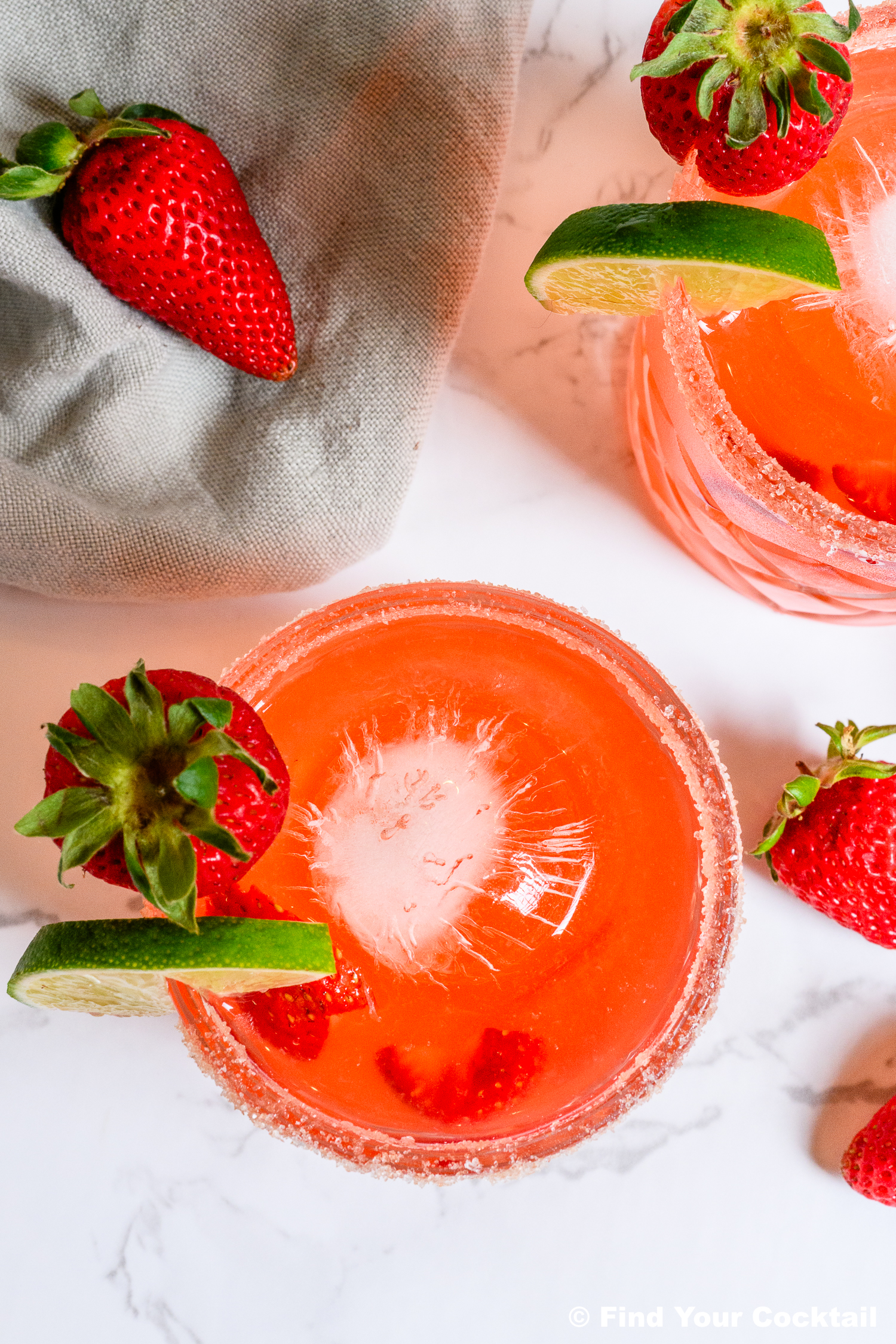 Overhead view of two glasses of pink cocktail with round ice, garnished with strawberries and lime slices, next to a beige napkin and loose strawberries.