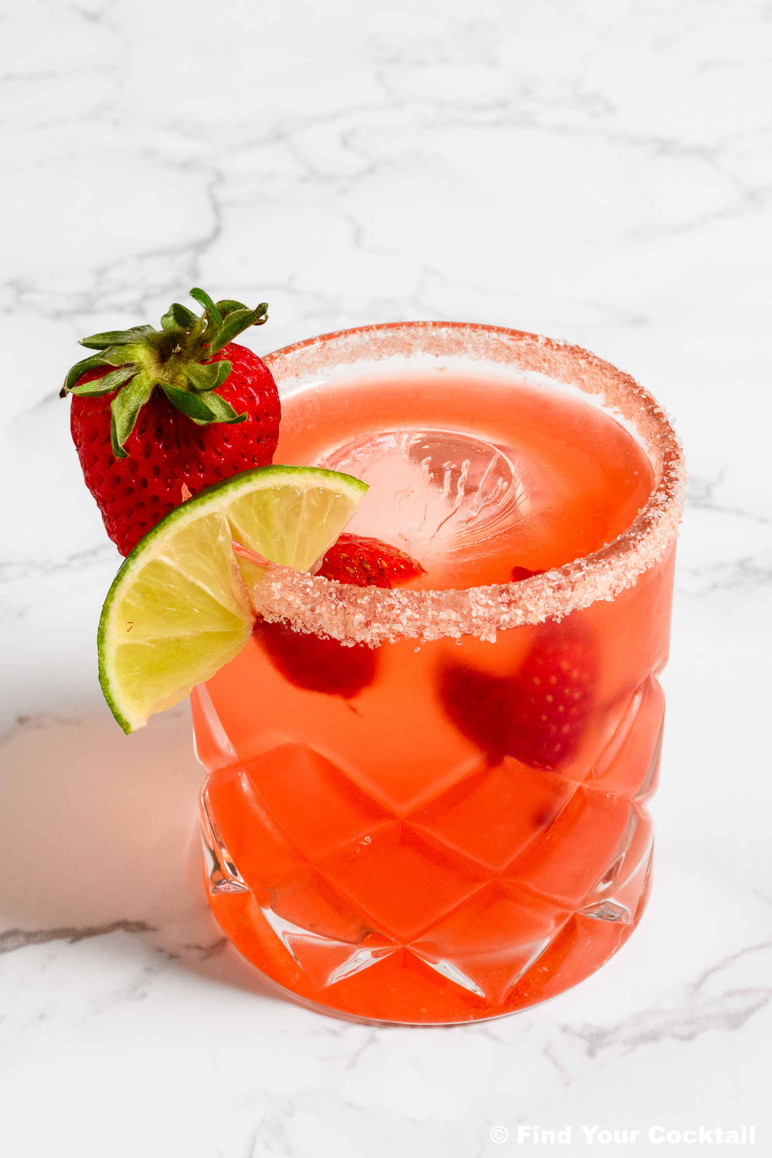 A glass of pink cocktail with a sugared rim, garnished with a fresh strawberry and lime slice, on a white marble surface.