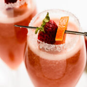 Two glasses of pink cocktail with sugared rims, garnished with a strawberry and orange slice on a cocktail pick.