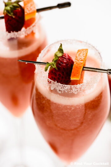Two glasses of pink cocktail with sugared rims, garnished with a strawberry and orange slice on a cocktail pick.