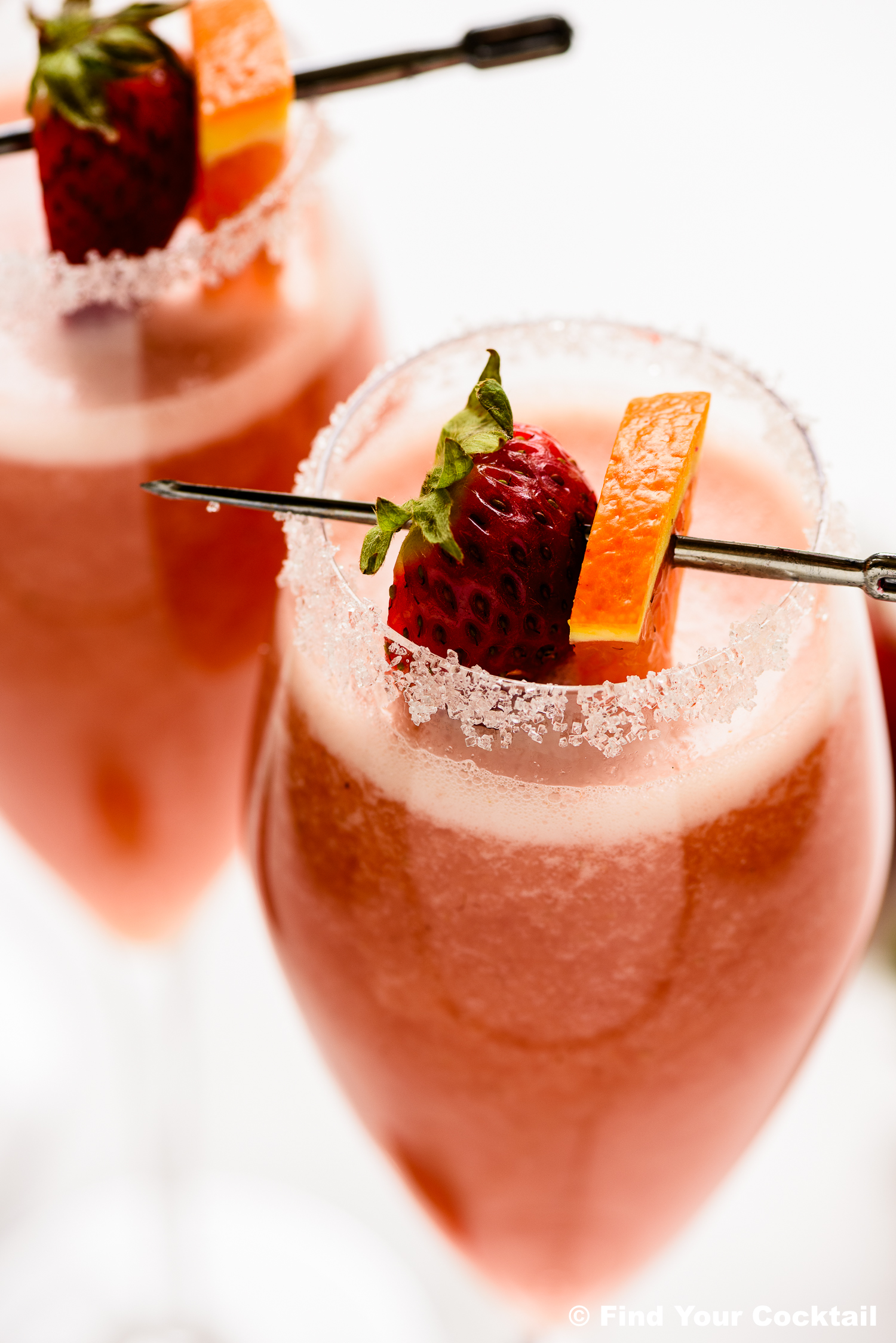 Two glasses of pink cocktail with sugared rims, garnished with a strawberry and orange slice on a cocktail pick.